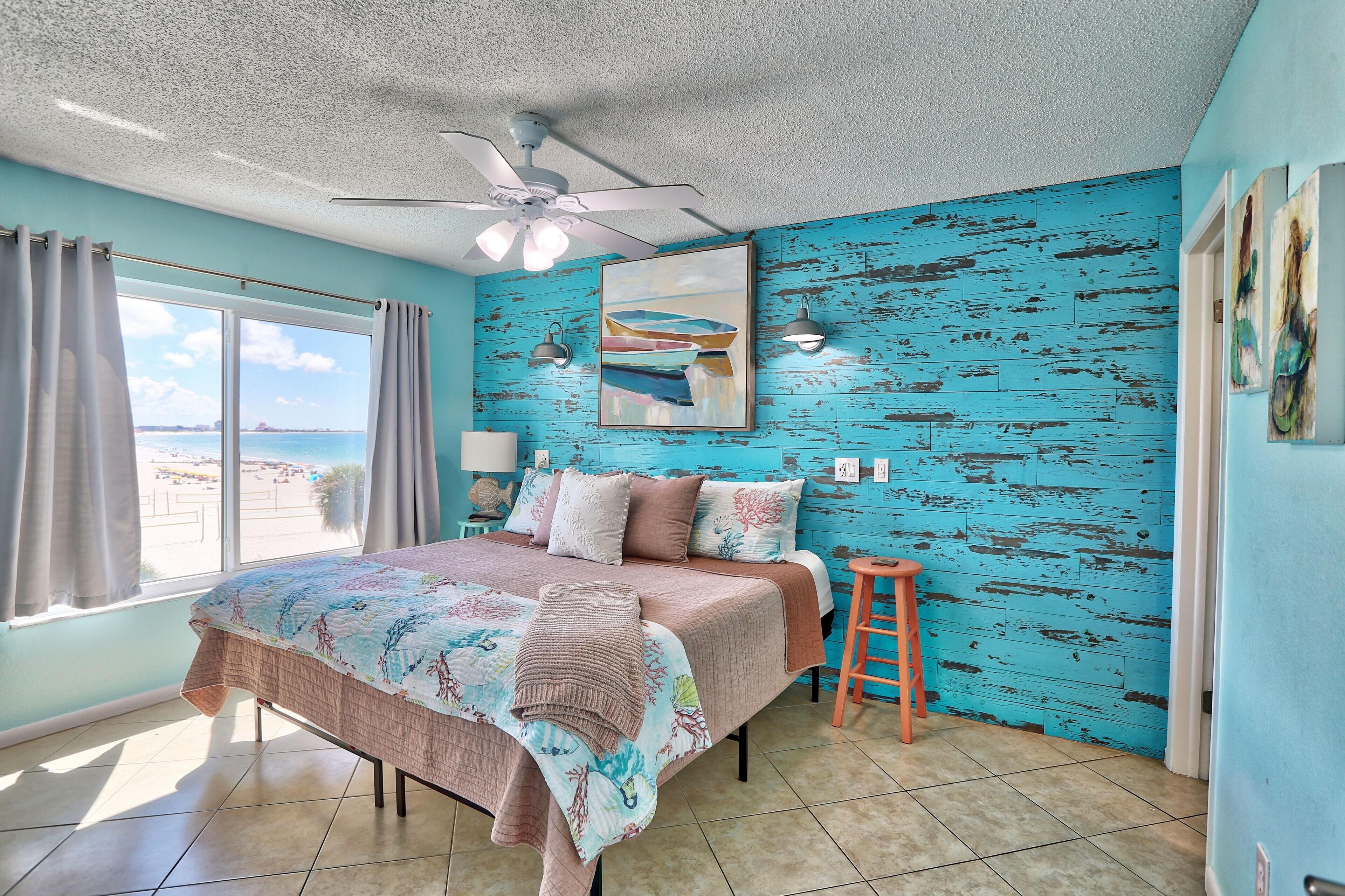 Un dormitorio con cama, ventilador de techo, ventana con cortinas, pared con acabado en pintura azul desgastada y un cuadro en la pared.