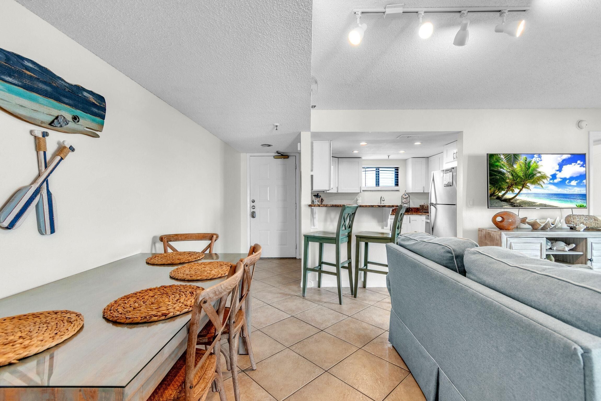 Una sala de estar moderna con un sofá gris, un comedor con una mesa de madera y individuales tejidos, y una cocina con armarios blancos y un televisor grande que muestra una escena de playa tropical.