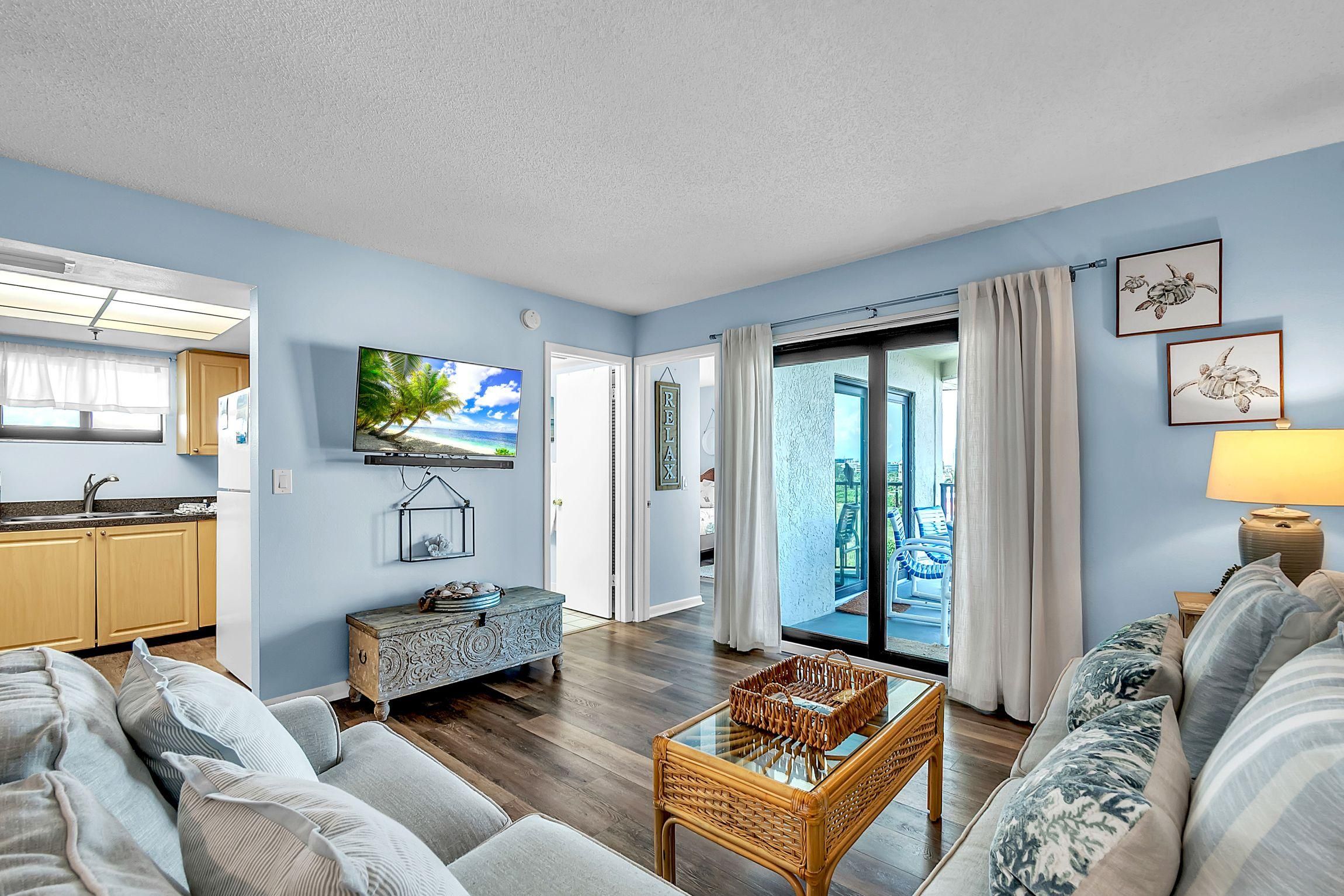 A living room with a sofa, a coffee table, and a television. There is a kitchen area with cabinets and a window.
