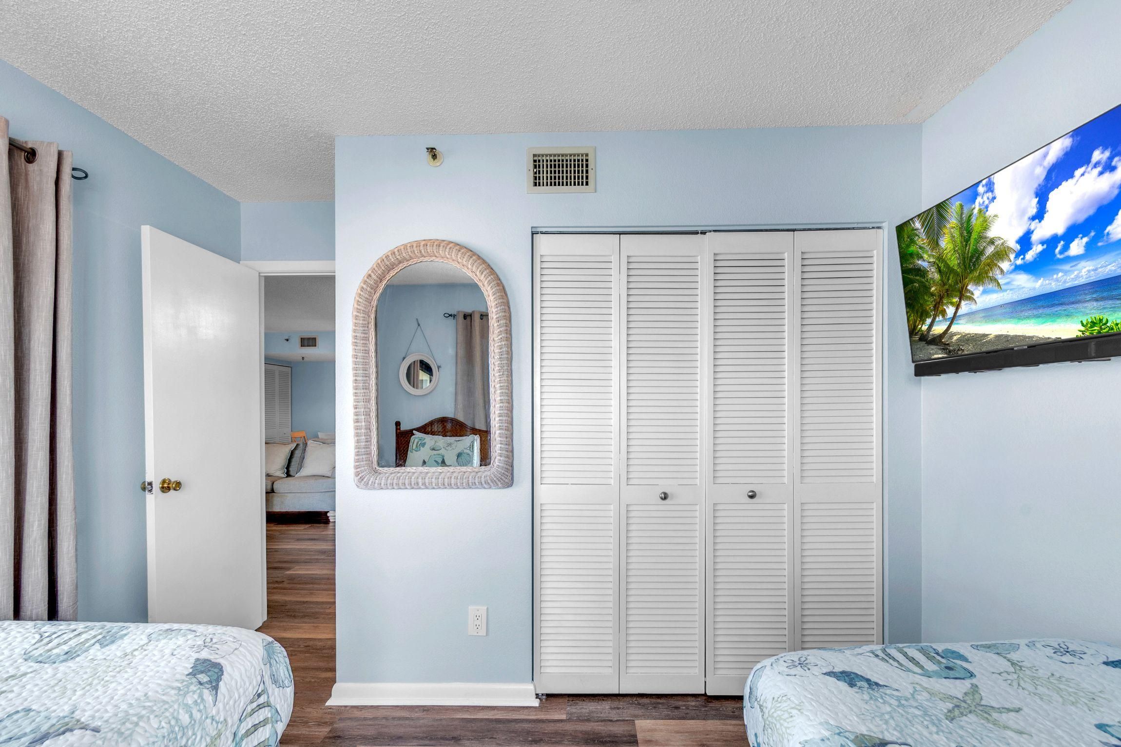 A bedroom with a bed, a wardrobe, a mirror, and a wall-mounted TV displaying a beach scene.