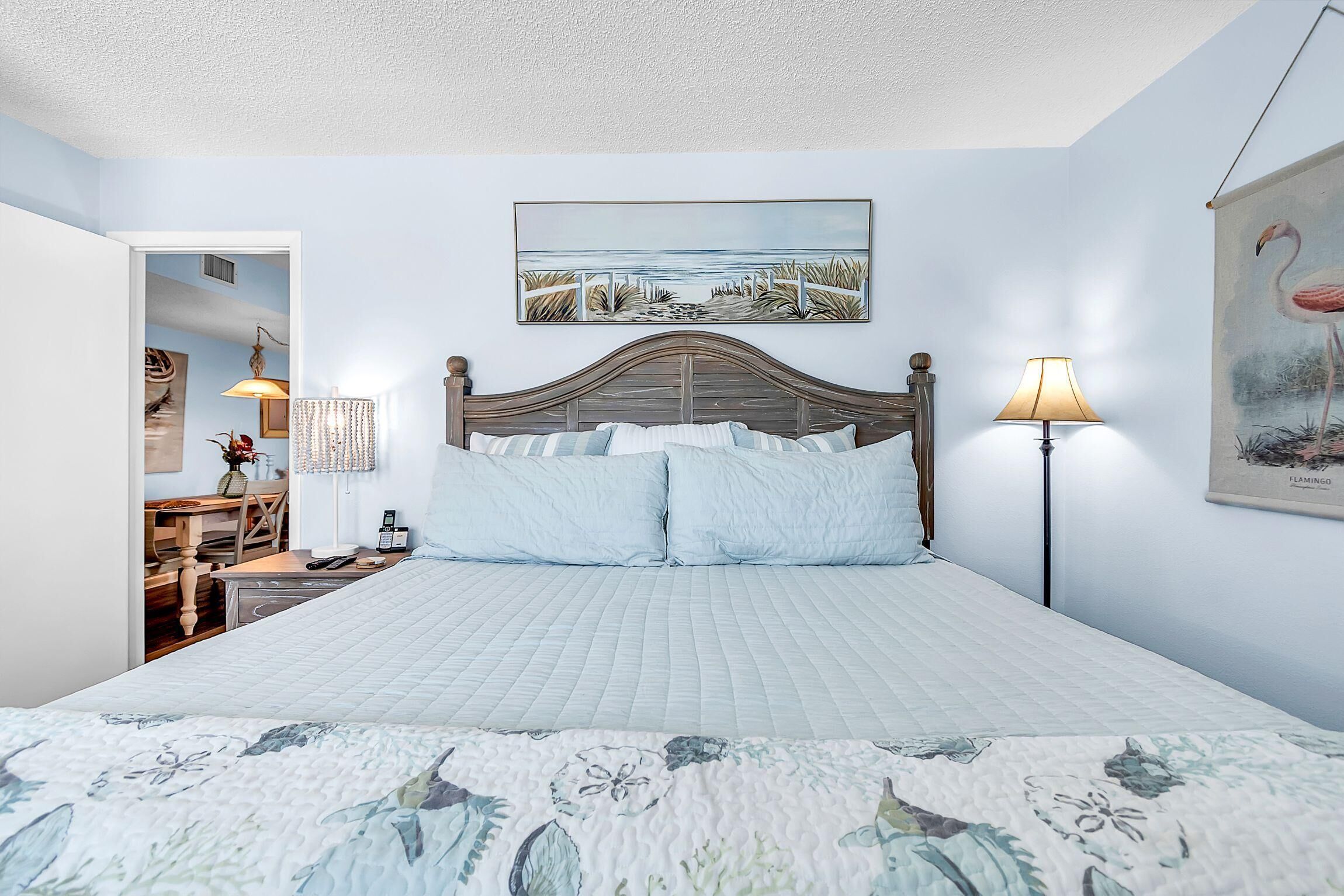 Een slaapkamer met een bed, een schilderij van een strand en een schilderij van een flamingo.