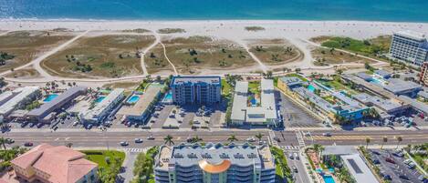 An aerial view of a coastal hotel complex with multiple buildings, a central structure with a distinctive roof, and a beach in the background.