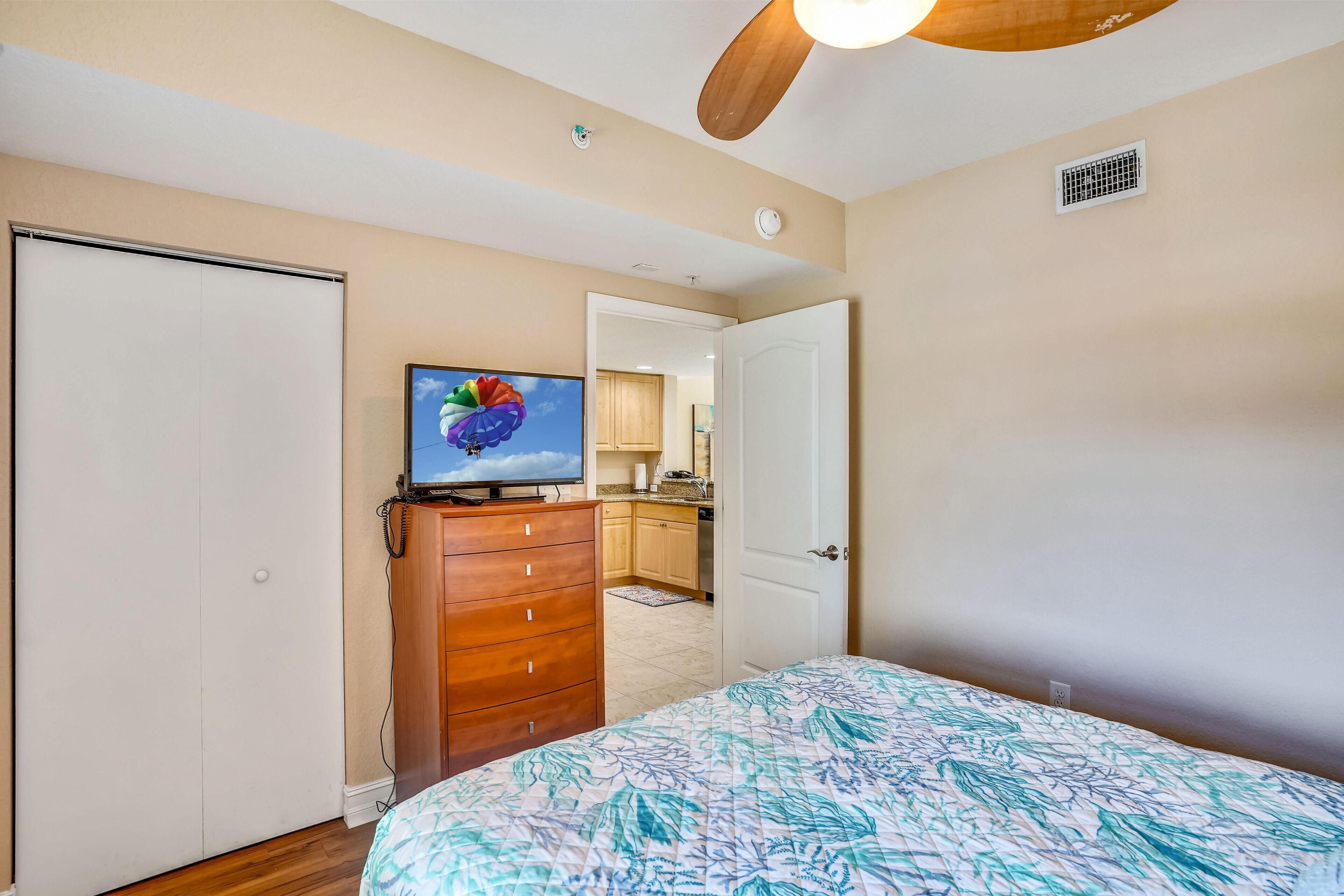 Een slaapkamer met een bed, een dressoir, een televisie en een deur die leidt naar een keuken.