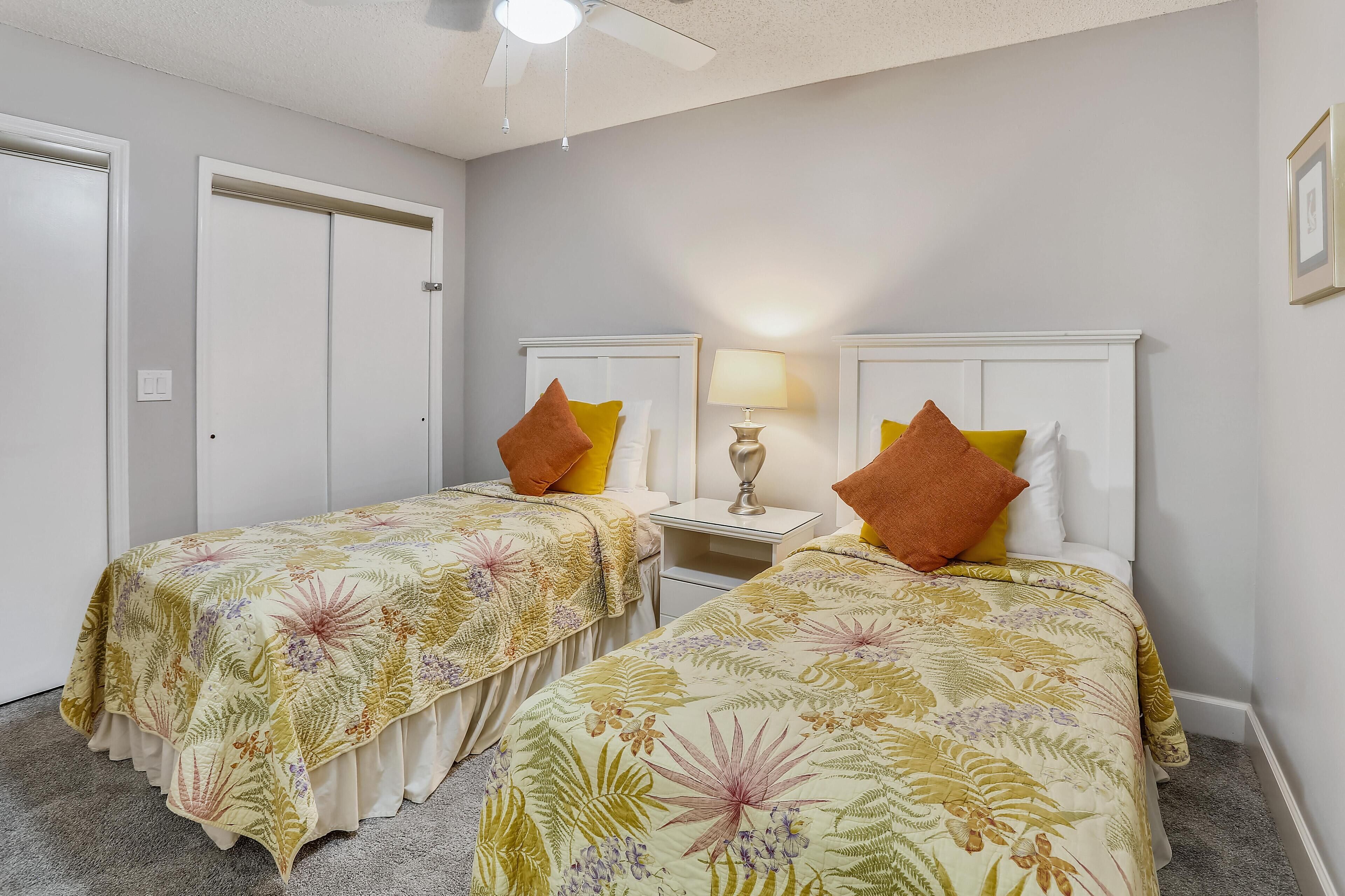 A hotel room with two beds, each with floral-patterned bedding, a bedside table with a lamp, and a ceiling fan.