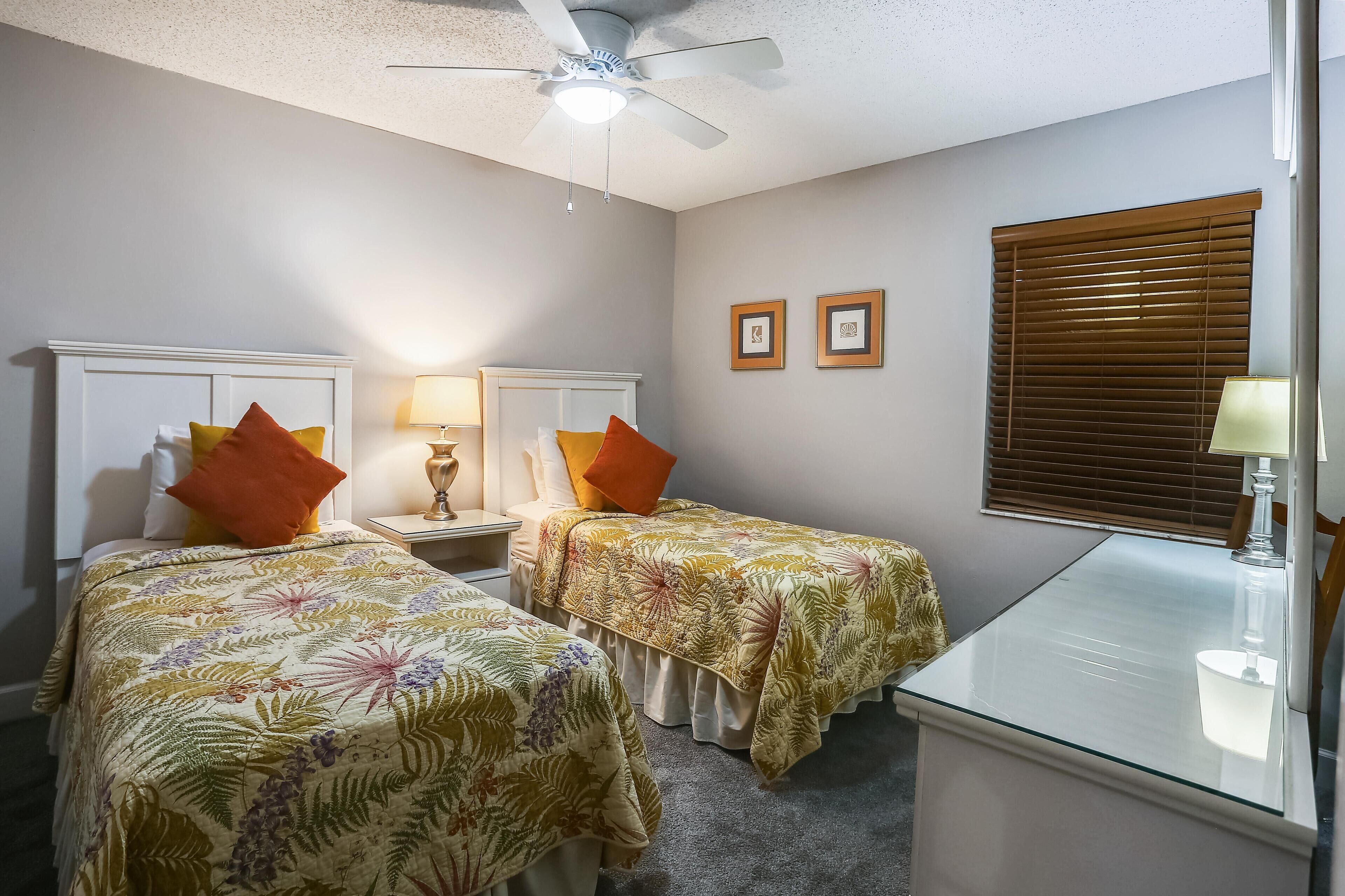 Une chambre d’hôtel avec deux lits simples, un ventilateur de plafond, un bureau et une fenêtre avec des stores.