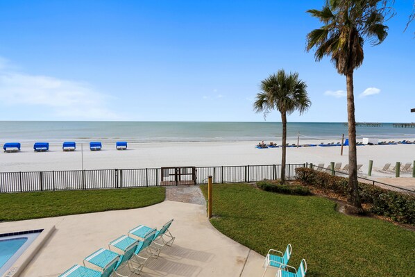 A pool area with lounge chairs, a palm tree, and a view of the beach with umbrellas and lounge chairs.