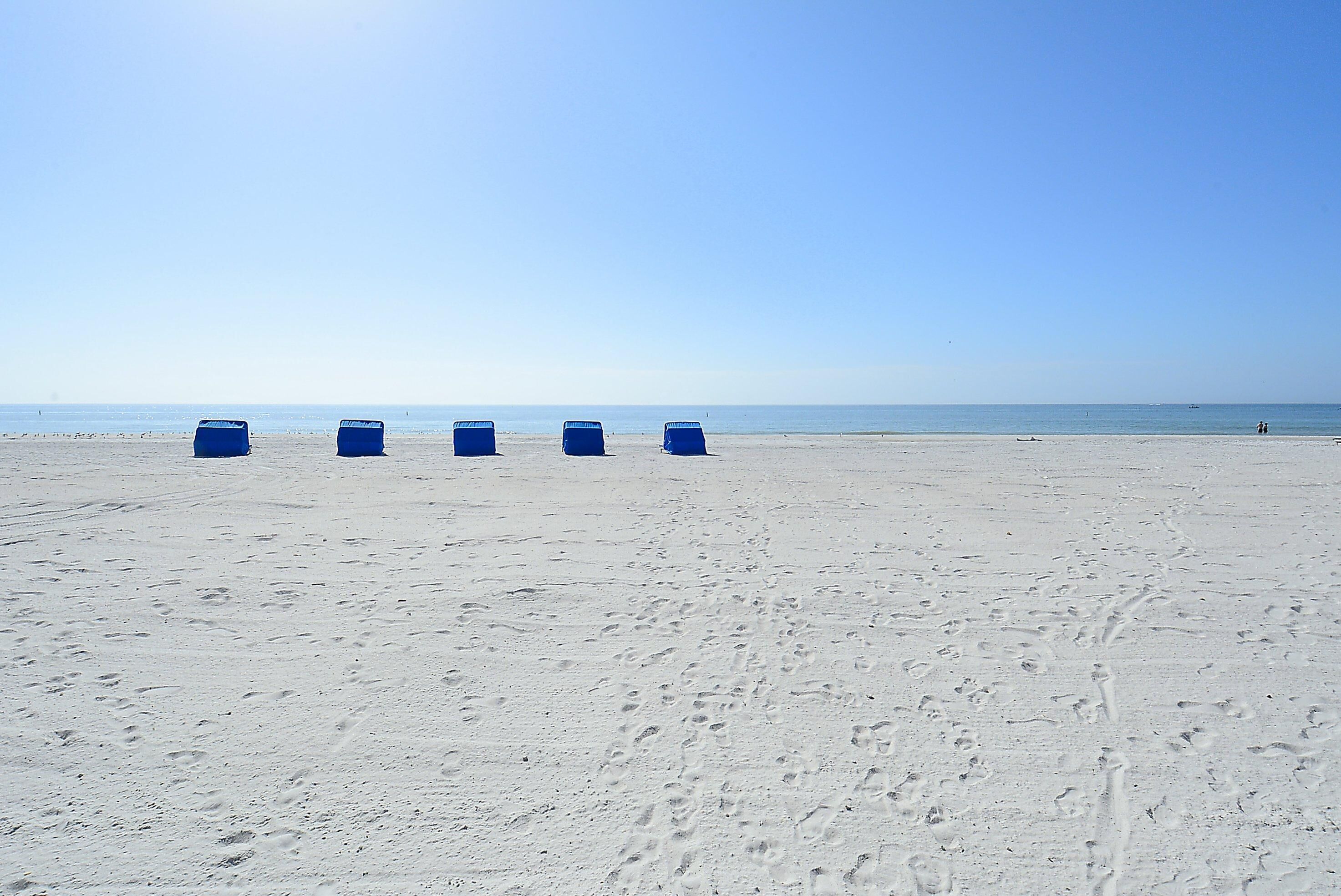 Una playa arenosa con cuatro botes de basura azules alineados.