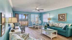 A living room with a teal wall, a ceiling fan, a sofa, a coffee table, and a dining area with chairs and a table. - Emerald Isle 104 (North Redington Beach)