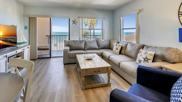 A living room with a sofa, coffee table, and a TV. There is a balcony with a view of the ocean. - Emerald Isle 204 (North Redington Beach)