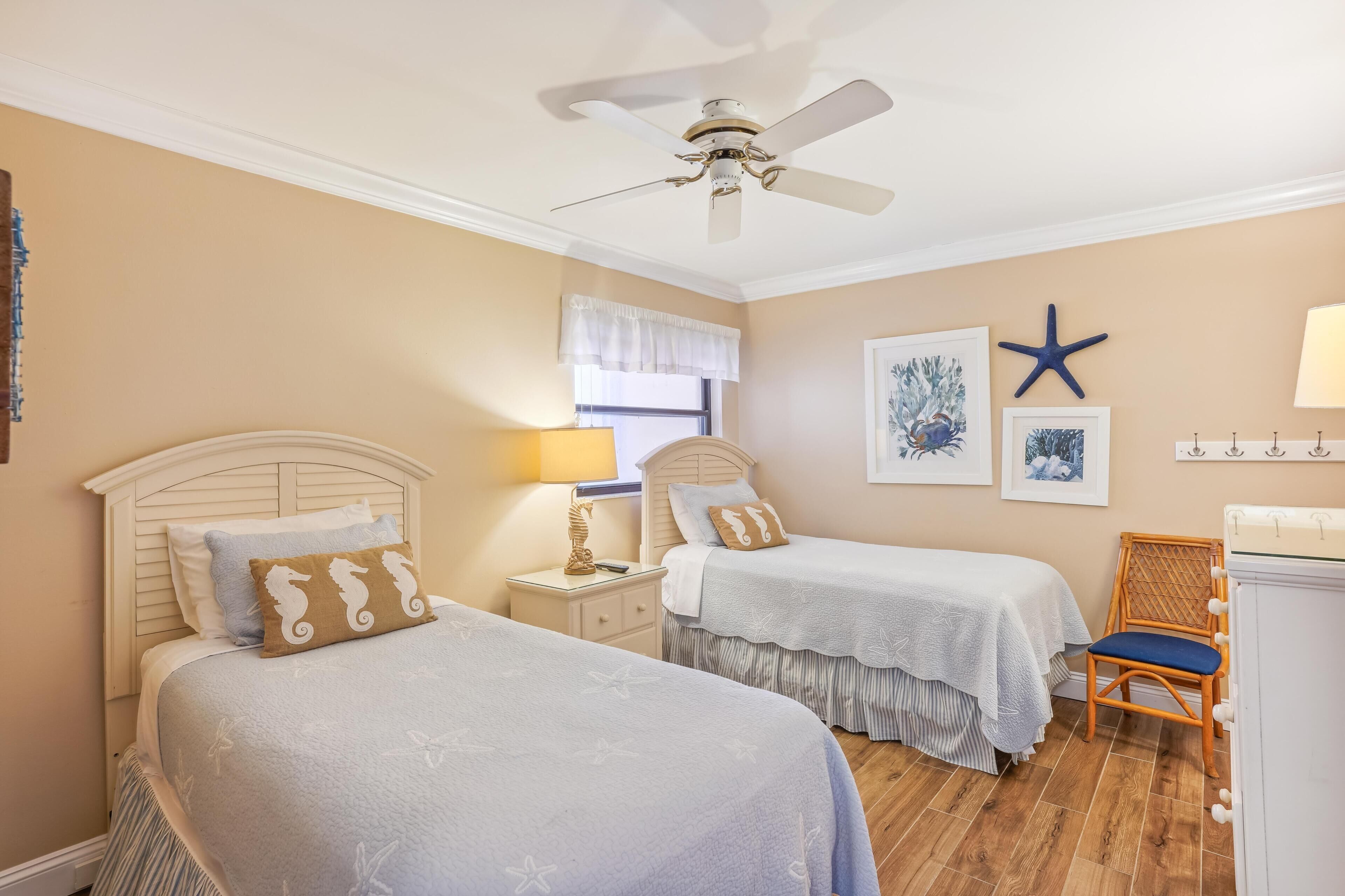 A bedroom with two beds, a ceiling fan, a window with a white valance, and a starfish decoration on the wall.