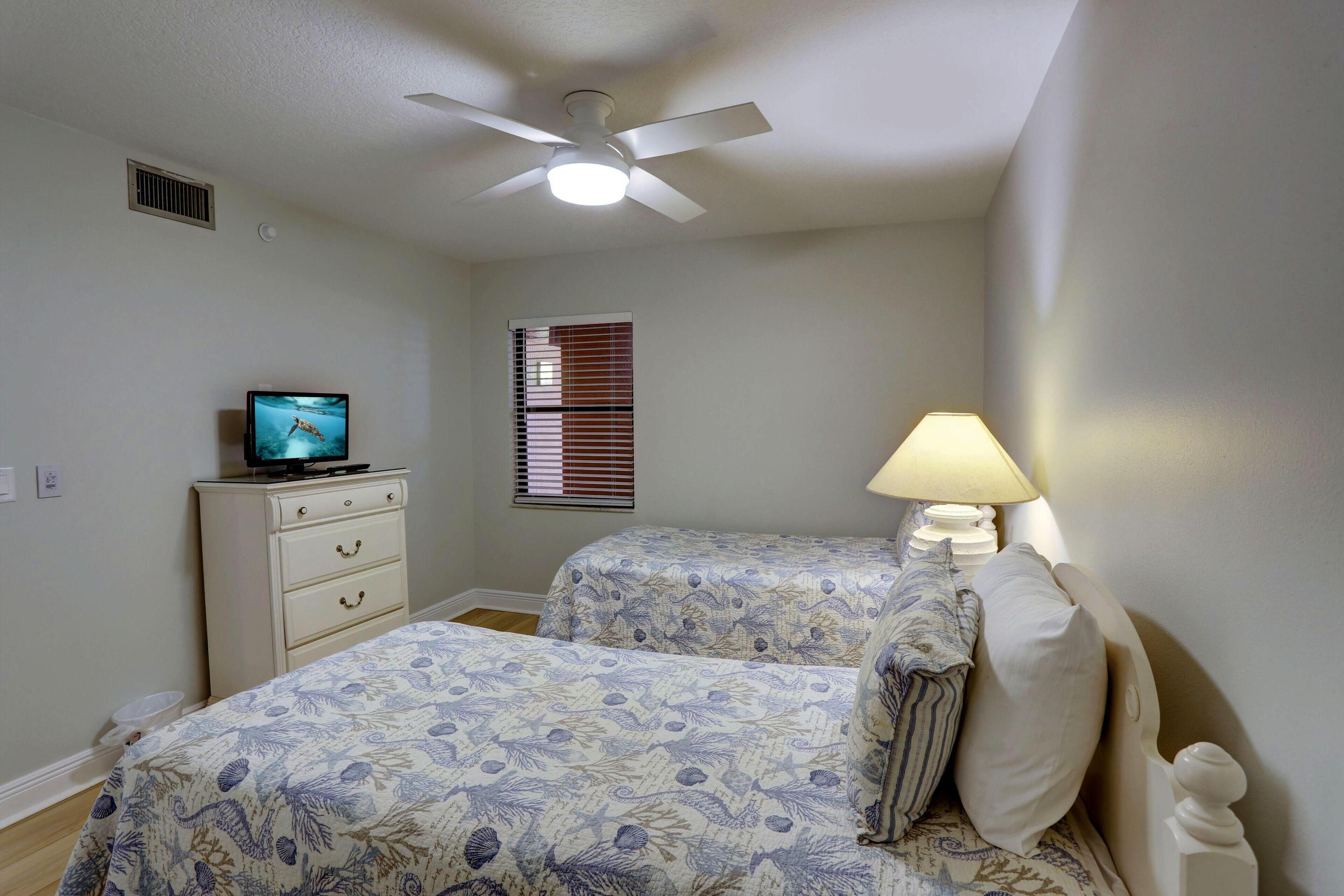 A bedroom with two beds, a ceiling fan, a TV on a dresser, and a window with blinds.