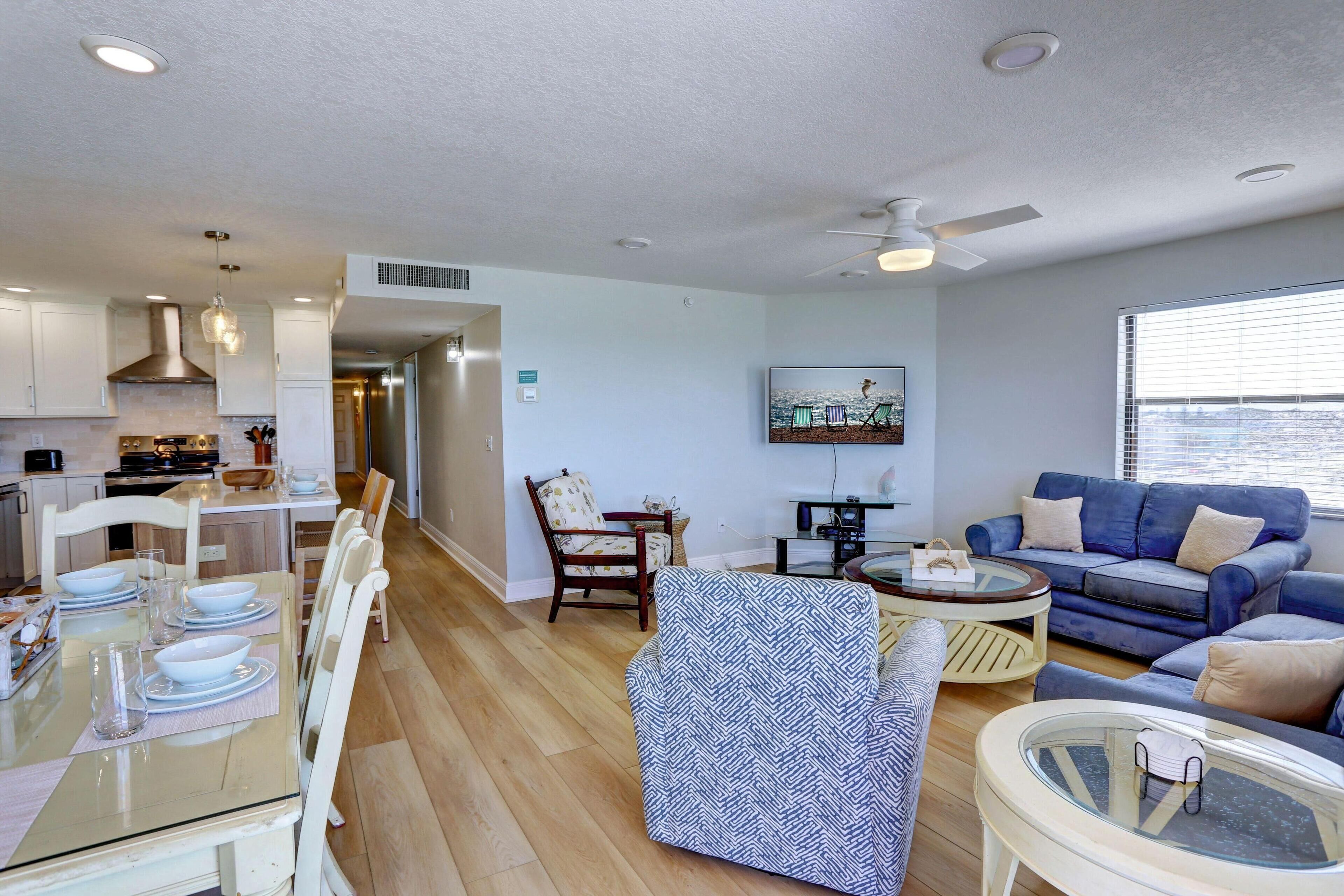 A modern living room with a blue sofa, a dining area with white chairs, and a kitchen with white cabinets.