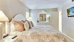 A bedroom with a bed, bedside tables, a dresser, a mirror, and a television. - Emerald Isle 601 (North Redington Beach)