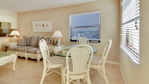 A living room with a glass-top coffee table, white wicker chairs, a striped sofa, and a dining table. - Emerald Isle 601 (North Redington Beach)