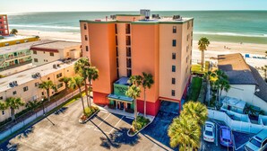 A multi-story hotel with a green and orange facade, located near a beach with a parking lot and several palm trees. - Emerald Isle 601 (North Redington Beach)