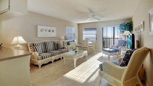 A spacious living room with a sofa, armchairs, and a coffee table. There is a ceiling fan, a dining area with chairs, and a kitchen area with a counter and a lamp. - Emerald Isle 601 (North Redington Beach)