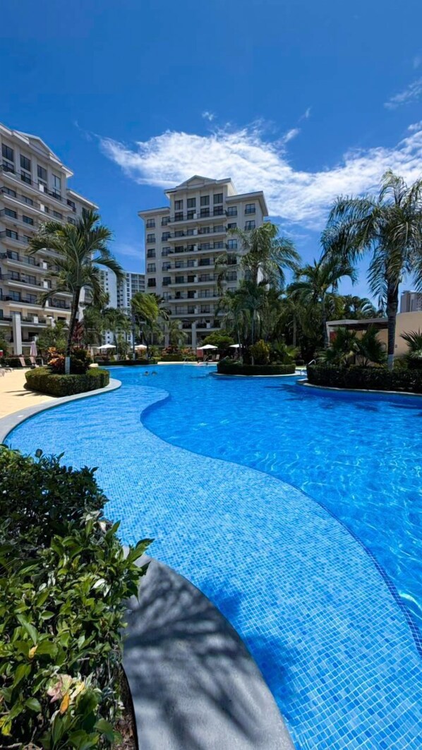 A swimming pool with a curved design, surrounded by palm trees and a building with multiple balconies.