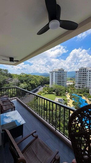 A balcony with a ceiling fan, a table with chairs, and a view of a resort with buildings and a pool.