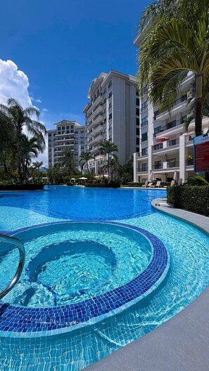 Une piscine dont le bord est recouvert de carreaux, entourée de palmiers et bordée d’un immeuble à plusieurs étages doté de balcons.