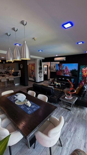 A modern living room with a dark wooden dining table, white cushioned chairs, a black sofa, and a TV. - Luxury Condo at Jaco Bay, Costa Rica's best beach (Jaco)
