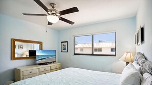 Une chambre à coucher avec un lit, un ventilateur de plafond, un téléviseur posé sur une commode, une fenêtre donnant sur un beau paysage et une lampe.