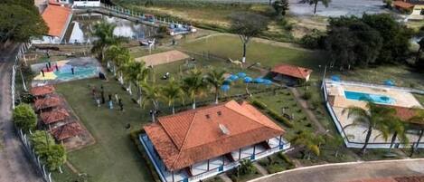 An aerial view of a resort with a swimming pool, a covered area with a red roof, and a fenced-in grassy area with palm trees.