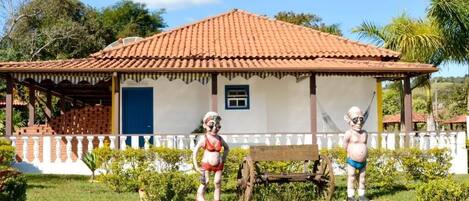 Una casa con techo de tejas, puerta azul y una ventana pequeña. En el patio delantero hay un banco y una estatua.