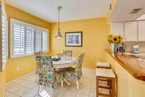 A dining area with a round table, floral chairs, a hanging light fixture, and a kitchen counter with a sink and a vase of sunflowers. - Gulfside Villas 6 (Indian Rocks Beach)