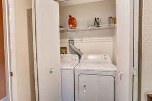 A laundry room with two white front-loading washing machines, a metal shelf above holding cleaning supplies, and a vent pipe running along the wall.