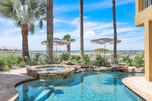 A swimming pool with a hot tub, surrounded by palm trees and umbrellas, with a view of the ocean.