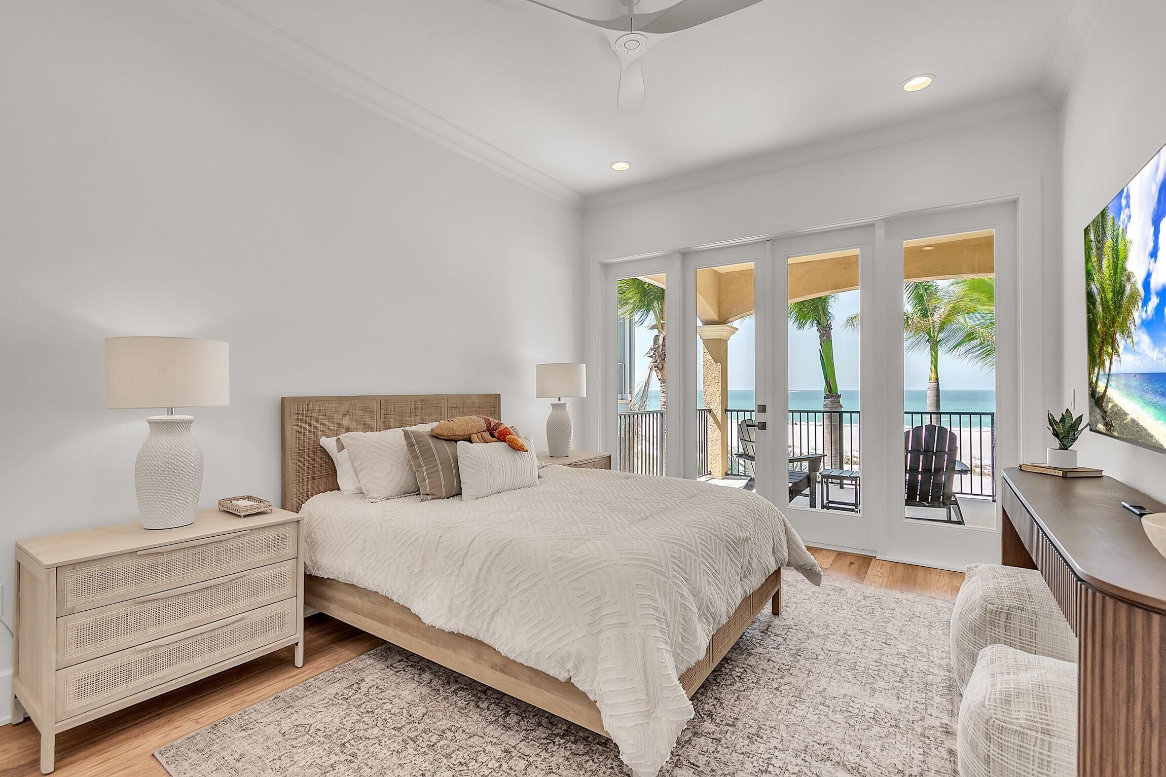 A bedroom with a bed, bedside tables, a dresser, a balcony with a view of the ocean, and a large window.