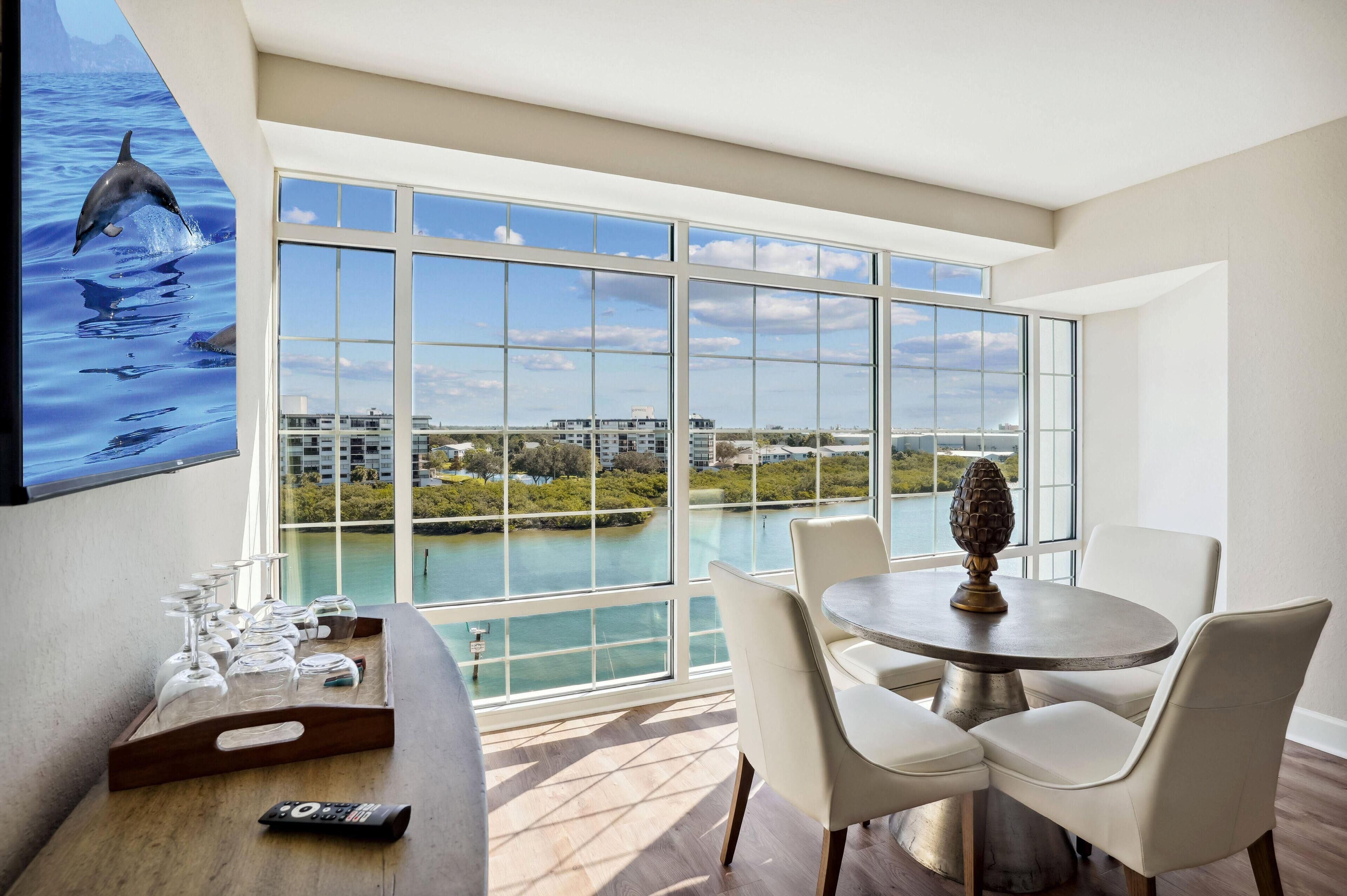 A dining area with a large window overlooking a body of water and buildings, a table with chairs, and a tray with glasses and a remote control.
