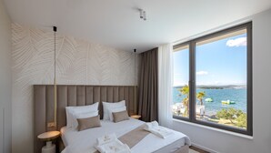 A modern bedroom with a large bed, a view of the sea, and a palm tree outside. - The Palms Resort - Villa President Beach I (Tkon)