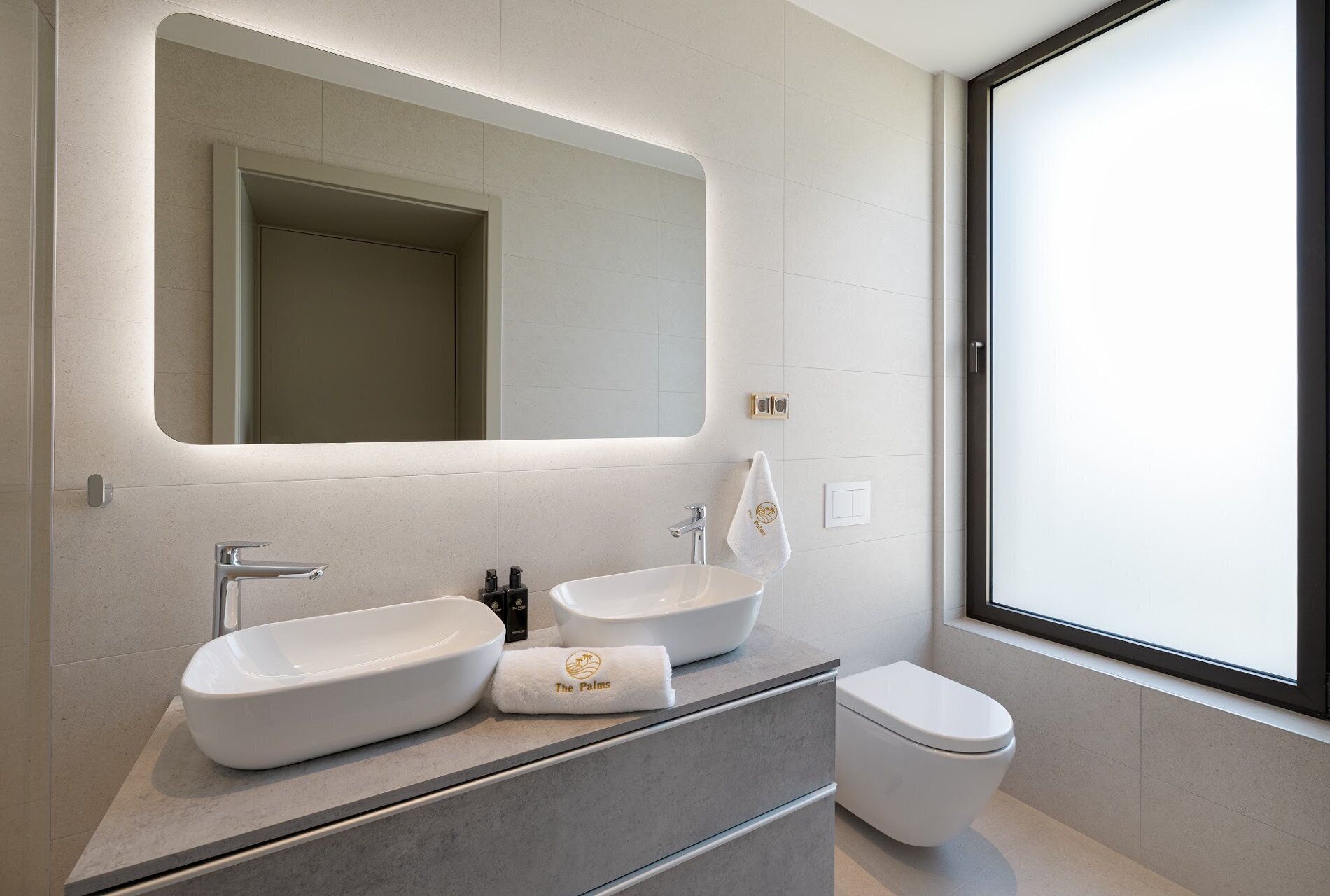 A modern bathroom with a large mirror, two sinks, a toilet, and a window.