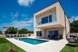 Ein modernes, zweistöckiges Haus mit Swimmingpool, Sitzgelegenheiten im Freien und einem Balkon.