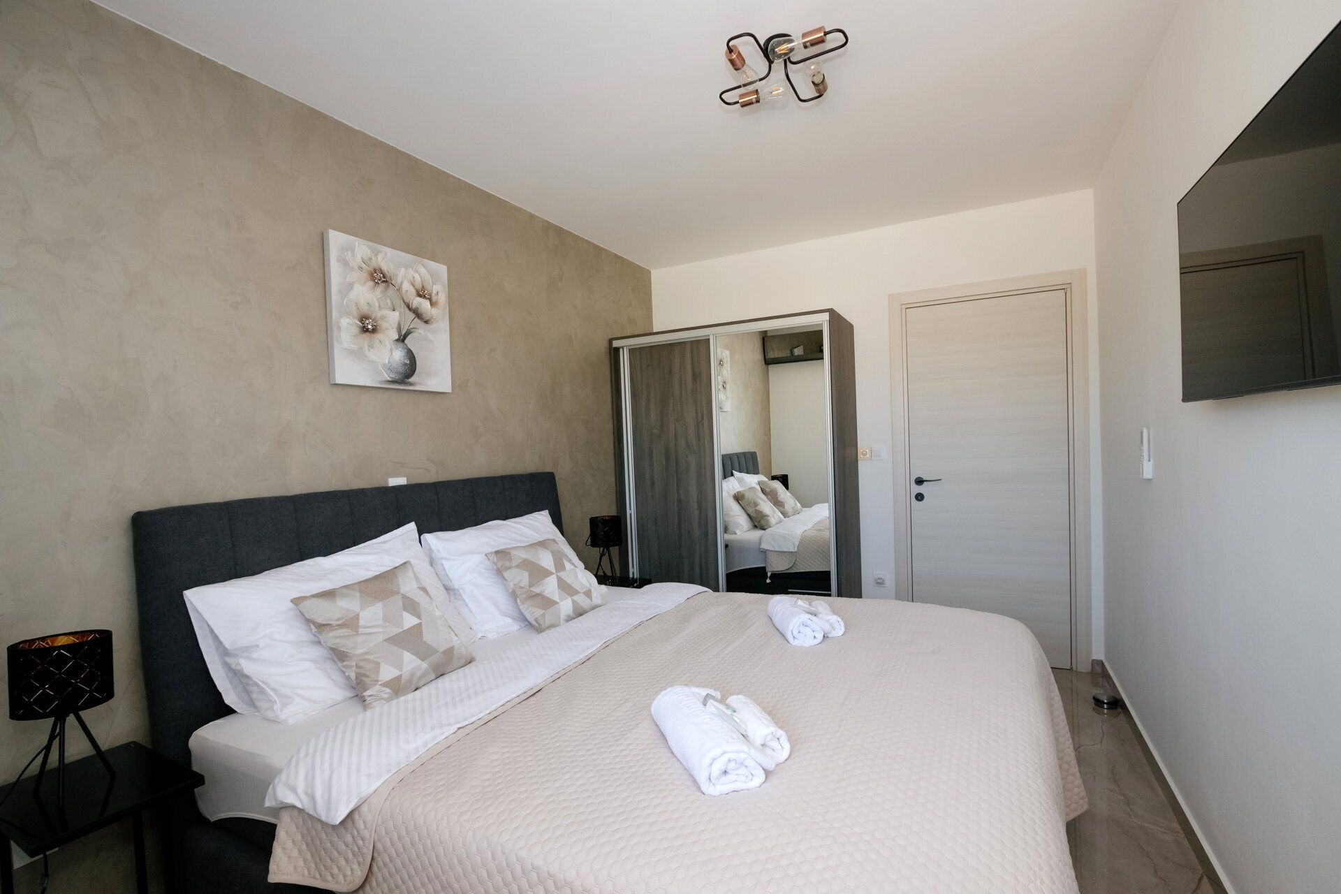 Habitación de hotel con una cama grande, una mesita de noche con lámpara, un espejo y televisión en la pared.