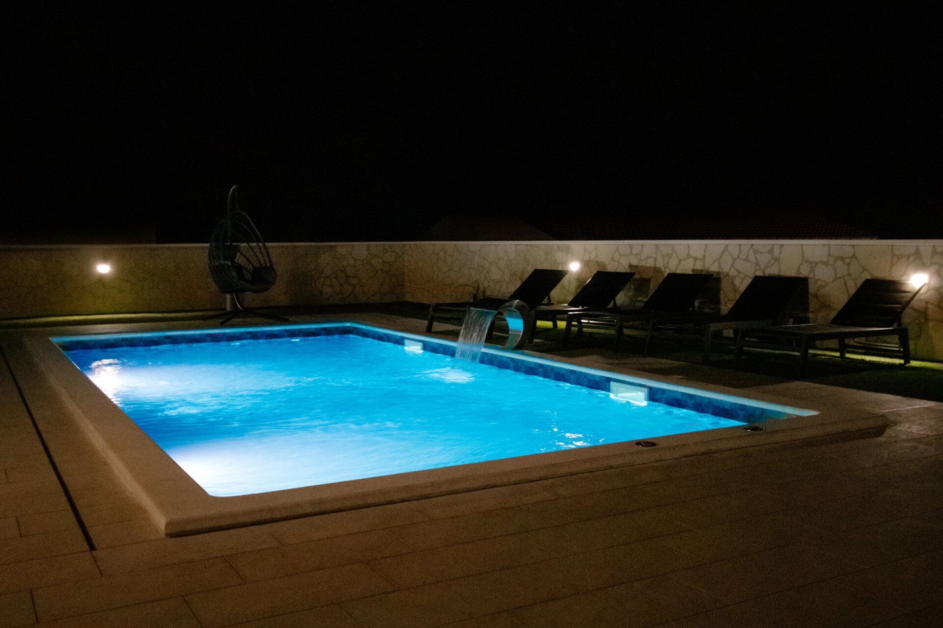 Una piscina rettangolare con elemento acquatico, circondata da sdraio e un muro con motivo in pietra.