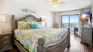 A bedroom with a large bed, a ceiling fan, a view of the ocean through a sliding door, and a television on a stand. - Las Brisas 202 (Madeira Beach)