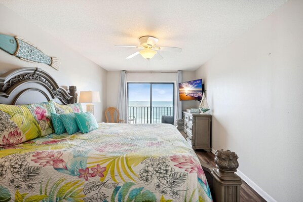 A bedroom with a bed, a ceiling fan, a TV, and a balcony with a view.