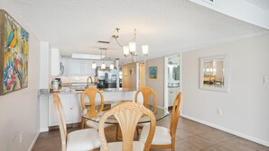 A dining area with a glass table and wooden chairs, a kitchen in the background, and a painting on the wall. - Las Brisas 202 (Madeira Beach)