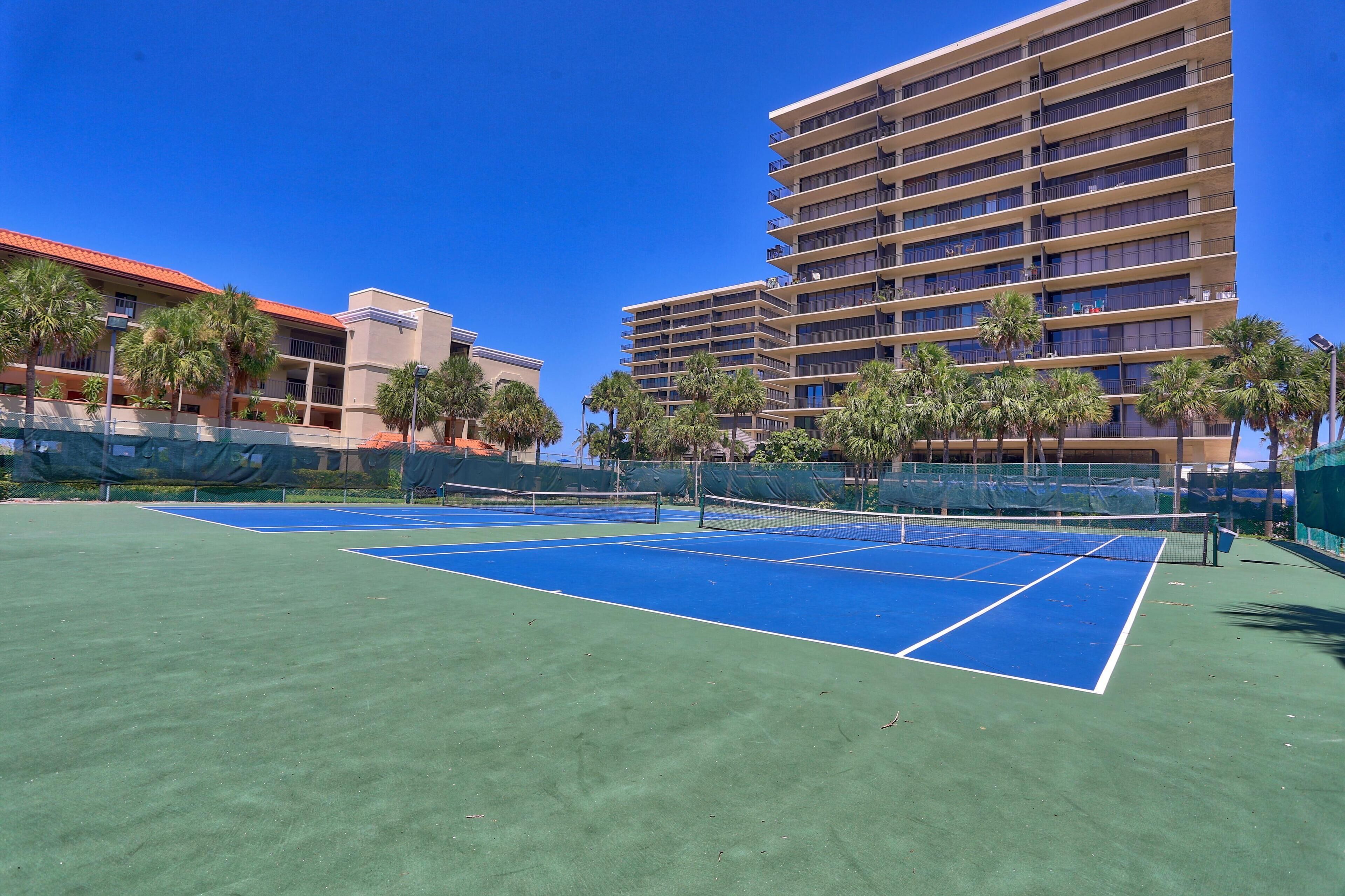 Una cancha de tenis con red, rodeada de palmeras y edificios.