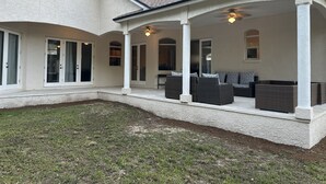 Een overdekt terras met wicker meubilair en plafondventilatoren, een witte gevel en een gazon.