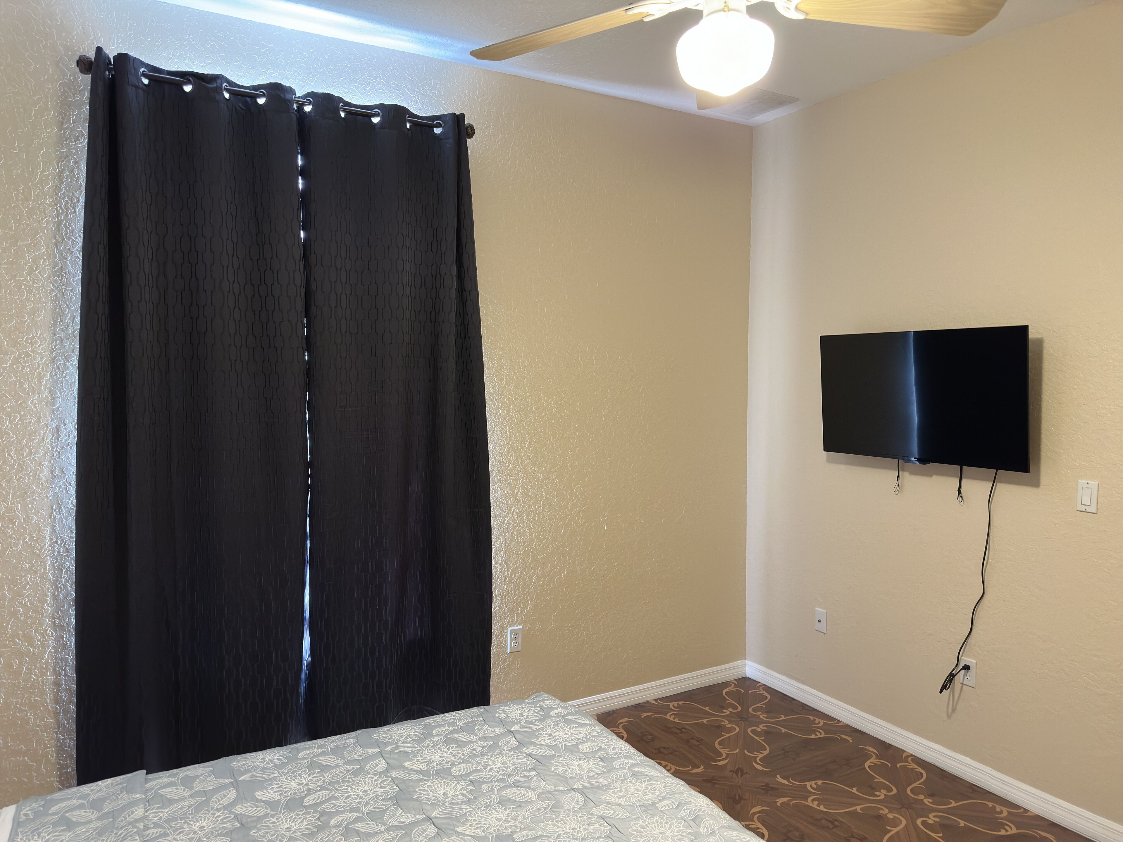 Un dormitorio con cama, ventilador de techo, televisor montado en la pared y ventana con cortinas.