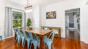 A dining area with a wooden table and light blue chairs, a hanging light fixture, a painting on the wall, and a view of the kitchen through an open door.