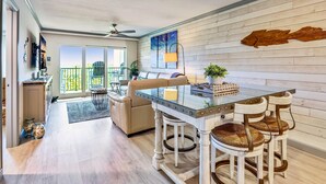 A modern kitchen with a glass-top table, white chairs, and a wooden wall with a fish decoration. - Land's End 10-202 Beach Front - Premier (Treasure Island)