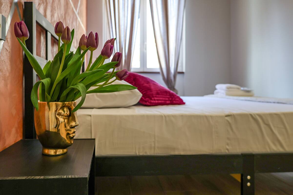 A bed with white bedding, a red pillow, and a vase with purple flowers on a black nightstand.