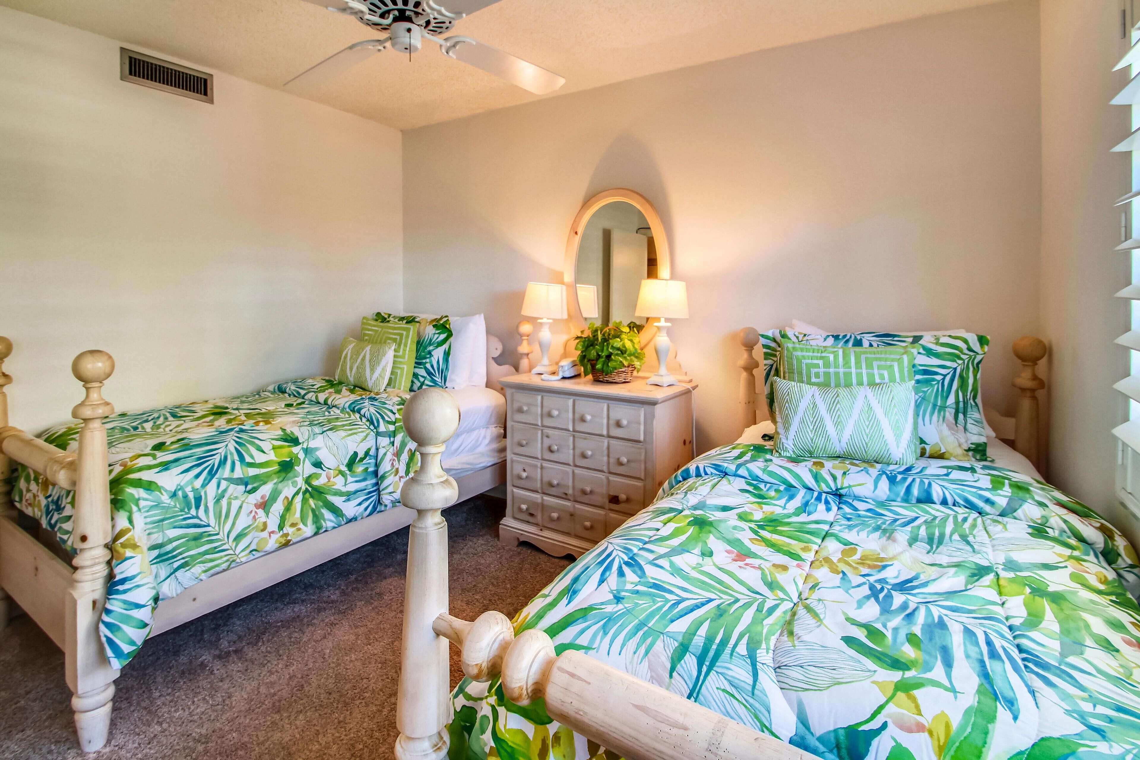 A bedroom with two wooden beds, each with a tropical leaf-patterned bedspread. There is a dresser with drawers and a mirror above it, and a ceiling fan above the beds.