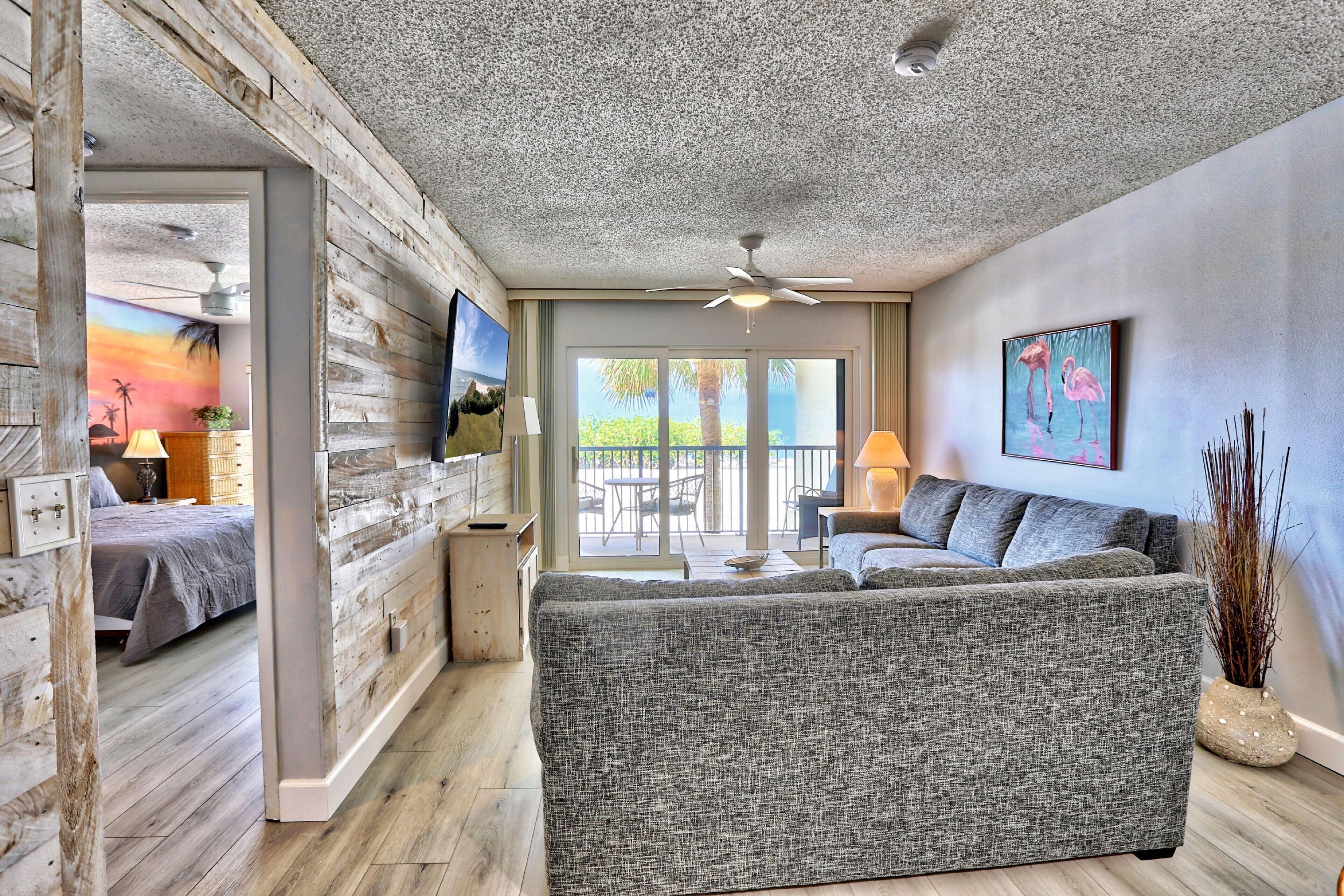 A modern living room with a grey sofa, wooden walls, a ceiling fan, and a large window overlooking a patio.