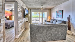 A modern living room with a grey sofa, wooden walls, a ceiling fan, and a large window overlooking a patio.