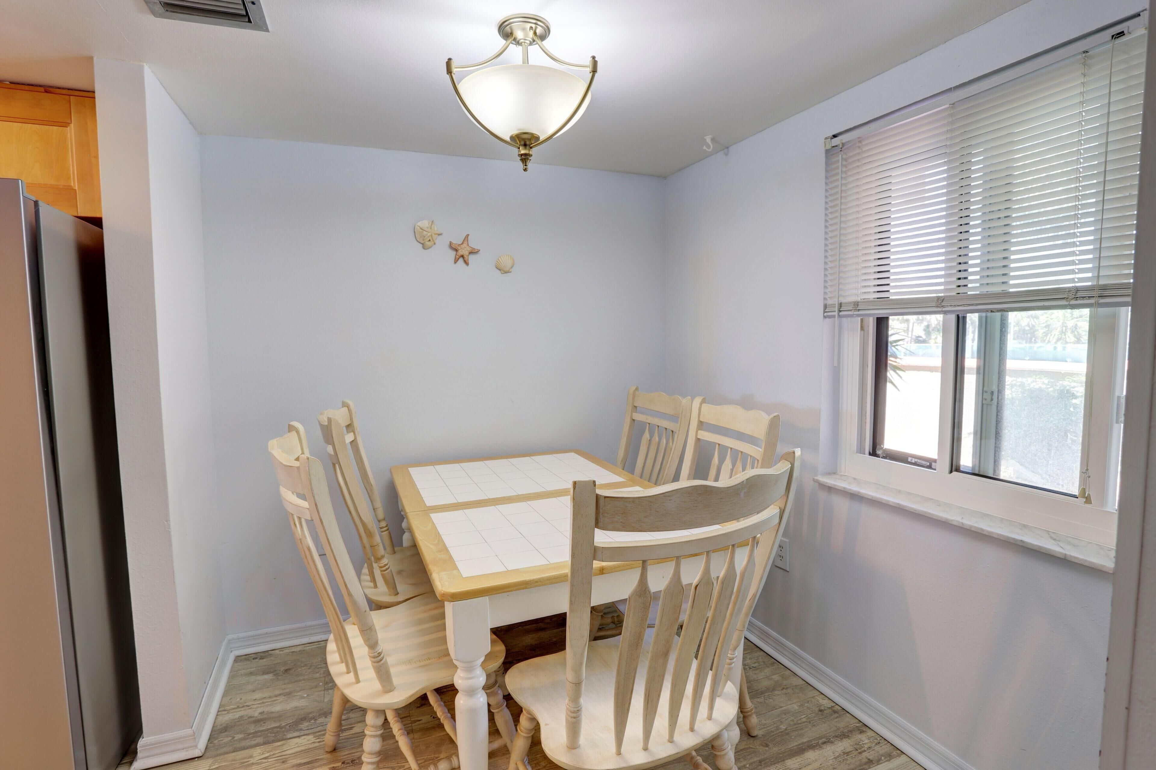 Un coin repas avec une table blanche et des chaises en bois, un luminaire au plafond et une fenêtre avec des stores.